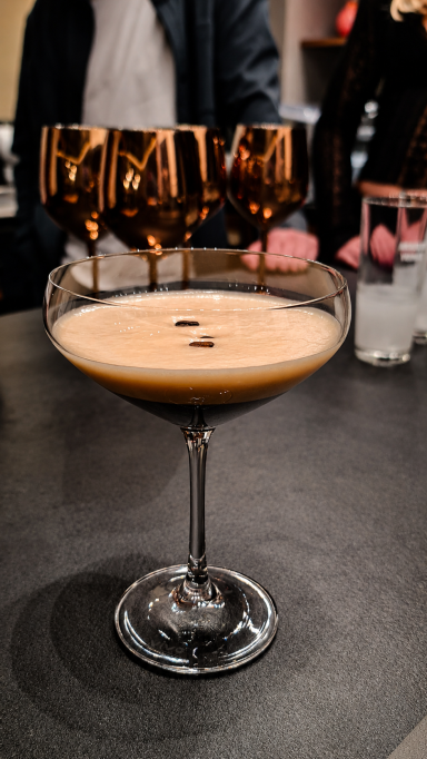 Cocktail in einer stilvollen Schale mit zwei Kaffeebohnen, im Hintergrund Gläser. Espresso-Martini