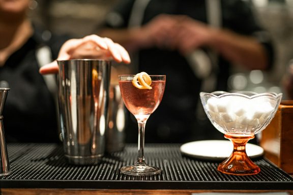 Ein Cocktailglas mit einem rosanen Getränk, neben einem Shaker und einer Schüssel mit Eis.