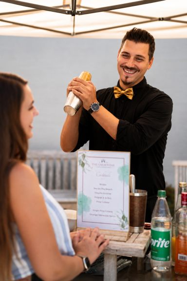 Barkeeper schüttelt einen Drink, während eine Kundin zuschaut. Getränke und Menü auf dem Tisch.